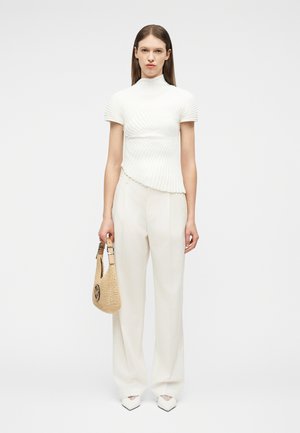 Mujer con una blusa blanca plisada de manga corta y pantalones de pierna ancha, sosteniendo un bolso tejido beige, posando frente a un fondo liso.