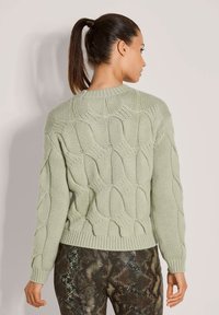 Frau mit braunem Pferdeschwanz, die einen hellgrünen, strukturierten Pullover und eine dunkelgemusterte Hose trägt, mit dem Rücken zu einer schlichten Hintergrund.