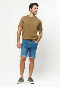 Mann steht mit der Hand in der Tasche, trägt ein braunes T-Shirt, hochgekrempelte Jeansshorts und grüne Turnschuhe vor weißem Hintergrund.