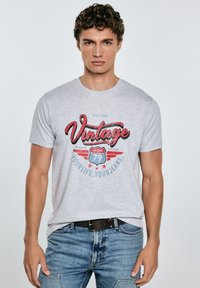 Pilkas medvilninis marškinėliai su "Vintage" grafika raudona ir mėlyna spalvomis, apvaliu kaklu, trumpomis rankovėmis, derinami su mėlynais džinsais ir tamsiu diržu.