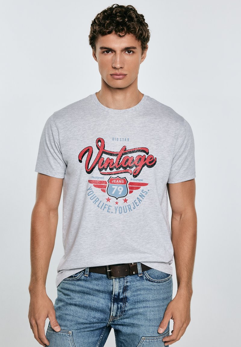 Pilkas medvilninis marškinėliai su "Vintage" grafika raudona ir mėlyna spalvomis, apvaliu kaklu, trumpomis rankovėmis, derinami su mėlynais džinsais ir tamsiu diržu.