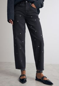 Schwarze Jeans aus Denim mit runden Ösen-Details, gerader Schnitt und hochgekrempelten Säumen, kombiniert mit schwarzen, spitzen Ballerinas mit Riemenakzenten.