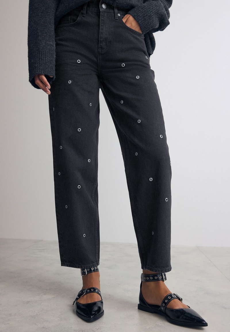 Schwarze Jeans aus Denim mit runden Ösen-Details, gerader Schnitt und hochgekrempelten Säumen, kombiniert mit schwarzen, spitzen Ballerinas mit Riemenakzenten.