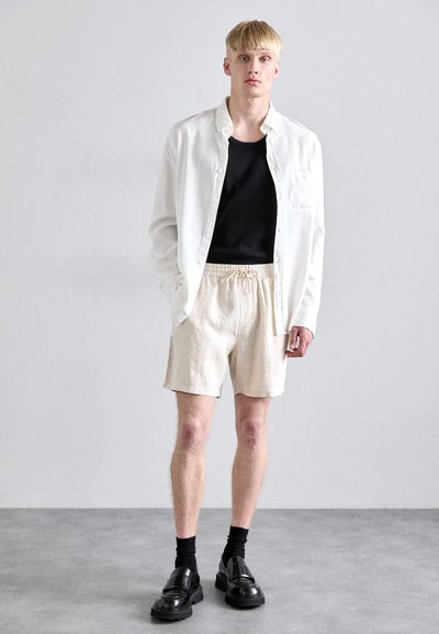 Chemise blanche à boutons sur un débardeur noir, associée à un short en lin beige et des chaussures épaisses noires. Design simple avec une taille élastique à cordon.