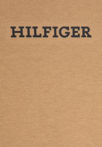T-shirt di cotone in una tonalità marrone chiaro con un prominente logo nero "HILFIGER". Il tessuto presenta una texture a coste.