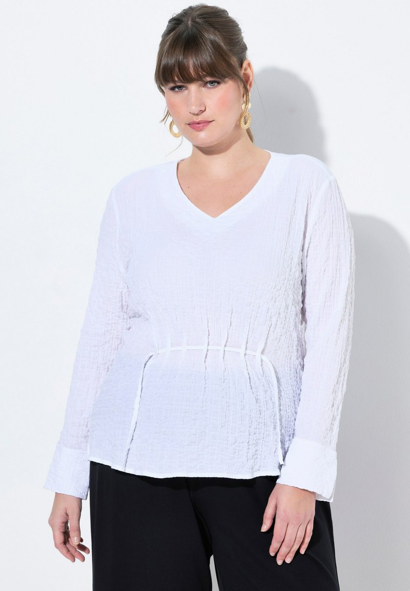 Valkoinen pitkähiha blouse, jossa on V-pääntie, teksturoitu kangas, kerätty vyötärö ja kaareva helma. Varustettu leveillä hihansuilla ja minimalistisella muotoilulla.