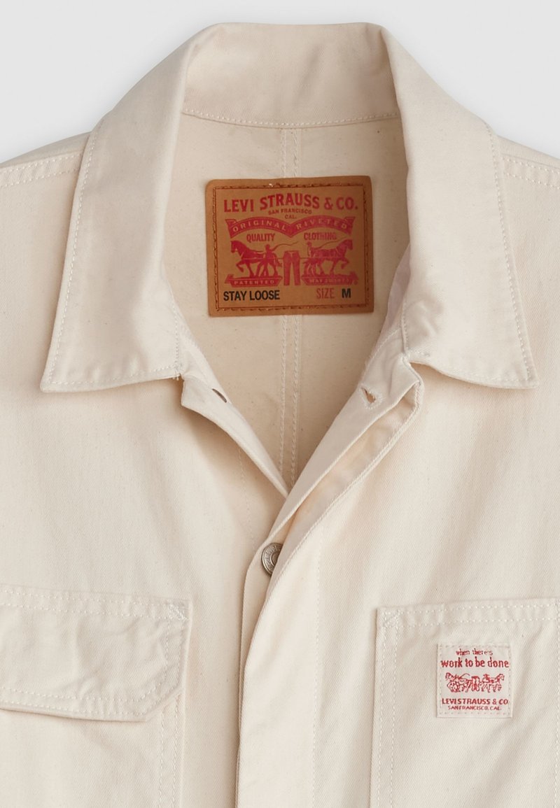 Chemise légère beige, à manches courtes, avec un col pointu, deux poches poitrine et une étiquette en cuir avec du texte rouge et des graphiques.