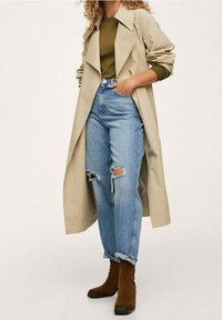 Trench-coat beige porté sur un haut vert olive, associé à un jean bleu clair délavé. Des bottines marron complètent la tenue.