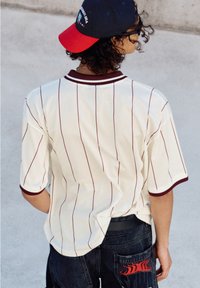 Weißes Mesh-Trikot mit kastanienbraunen Streifen, kurzen Ärmeln und geripptem Kragen. Getragen mit dunklen Jeans, die ein rotes Flammendesign an der Tasche haben.