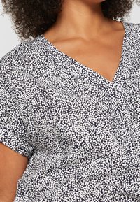 Blouse en polyester avec un fond bleu marine orné d'un motif à pois blancs, manches courtes et col en V. Tissu léger, texture lisse.