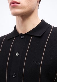 Κοντινό πλάνο ενός ατόμου που φοράει μαύρο πουκάμισο με γιακά, με λεπτές μπεζ κάθετες ρίγες και τρία μαύρα κουμπιά.