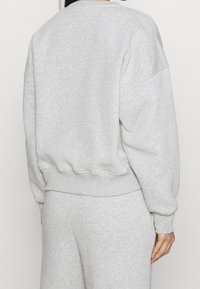 Persona vistiendo un sudadera gris claro y pantalones deportivos a juego, mostrada de espaldas contra un fondo liso.