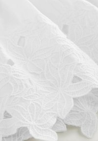 Tissu blanc brodé présentant des motifs floraux complexes avec des découpes et une surface texturée. Les bords sont festonnés.