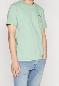 T-shirt en coton vert menthe avec un col rond, des manches courtes et un petit logo bleu marine sur la poitrine gauche. Porté avec un jean bleu clair.