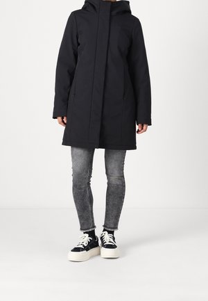 Un mannequin porte un manteau noir à capuche, un jean gris et des baskets noires à plateforme sur un fond blanc uni.