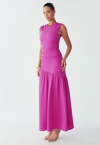 A woman stands confidently in a vibrant pink, fitted dress with a flared skirt, accessorized with subtle jewelry and open-toed shoes.