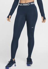 Leggings Nike Pro blu navy con fascia in vita bianca con logo, design aderente, texture liscia, logo Nike visibile sulla gamba inferiore, abbinati a scarpe da ginnastica.
