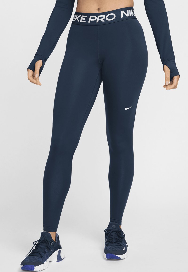 Leggings Nike Pro blu navy con fascia in vita bianca con logo, design aderente, texture liscia, logo Nike visibile sulla gamba inferiore, abbinati a scarpe da ginnastica.