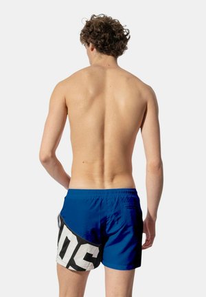 Giovane uomo con capelli ricci castani visto di spalle, indossa un costume da bagno blu con una grande grafica bianca e nera sul lato.