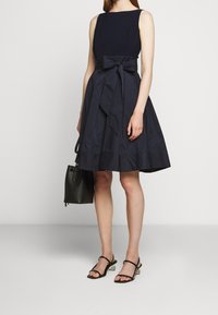 Nainen, jolla on yllään ilmanhihoja oleva tummansininen mekko, jossa on vyötäröllä solmu, mustat korkokengät ja musta käsilaukku, seisoo yksinkertaista taustaa vasten.