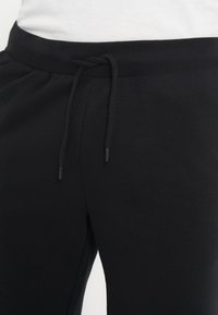 Pantalon de survêtement noir avec un cordon noué en nœud à la taille, porté avec un haut blanc visible à la taille.