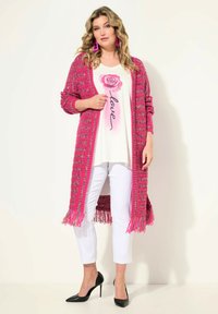 Długi różowy kardigan z frędzlami, o plecionej strukturze, noszony na białej koszuli z graficznym motywem róży i czarnym napisem, w połączeniu z białymi spodniami i czarnymi szpilkami.