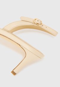 Beige High-Heelsandalen mit einem dünnen Riemen, einer runden Zehenpartie und einem goldenen Schnallen-Detail. Glatte Textur mit einem eleganten, minimalistischen Design.