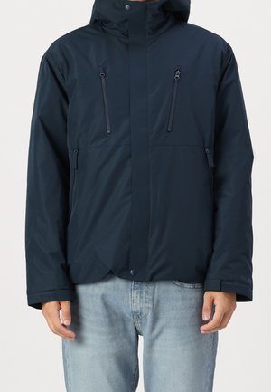 Homme portant une veste à capuche bleu marine avec quatre poches zippées et un jean bleu clair, debout devant un fond uni.