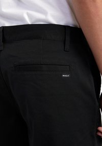 Pantalones negros de algodón con una textura suave. Cuenta con un bolsillo trasero y una pequeña etiqueta de logo de RVCA. El diseño es simple y contemporáneo.