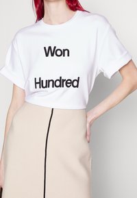 Camiseta blanca de manga corta con texto negro que dice "Ganado" y "Cien". La camiseta tiene un corte relajado y mangas enrolladas, combinada con una falda beige que presenta una costura negra.
