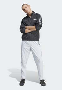 Giacca a vento nera con strisce bianche, abbinata a pantaloni della tuta grigi chiari. Sneaker scure completano il look sportivo.