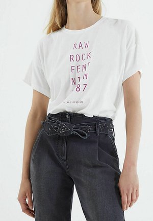 Kobieta w białym t-shircie z czerwonym napisem i ciemnych jeansach z wysokim stanem i paskiem z ćwiekami zawiązanym w kokardę.