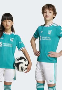 Dvoje djece nose akva nogometne dresove s bijelim detaljima i logotipom Liverpoola. Jedno drži crno-bijelu nogometnu loptu. Bijele kratke hlače i čarape.