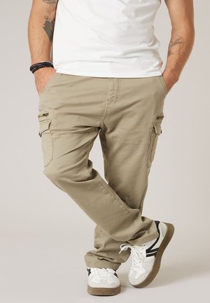 Homme portant un pantalon cargo beige, des baskets blanches avec des accents noirs, un t-shirt blanc, des tatouages sur les bras et des bracelets noirs, debout avec les jambes croisées.