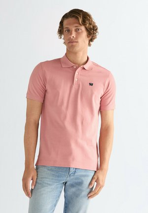 Polo rosa claro de manga corta, con cuello y un pequeño logotipo negro en el pecho, combinado con jeans azul claro.