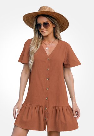 Kobieta ubrana w rdzawobrązową sukienkę z przodu zapinaną na guziki, z krótkimi, falbankowymi rękawami, dużym słomkowym kapeluszem, okularami przeciwsłonecznymi i złotą biżuterią.