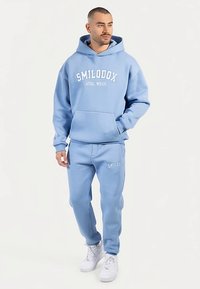 Ljusblå hoodie och matchande mjukisbyxor i mjukt tyg, med texten "SMILODOX ATHL WEAR" i vitt. Inkluderar känguruficka.