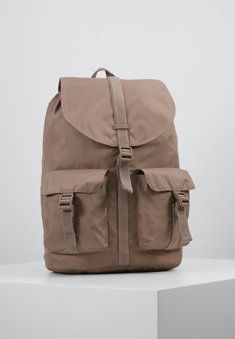 Sac à dos en tissu marron avec rabat supérieur et fermeture à boucle, doté de deux poches avant avec sangles à boucle, placé sur une surface blanche.
