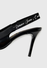 Black patent high-heeled sandal with a glossy finish, featuring a wide adjustable black strap with "Versace Jeans Couture" in white script lettering.