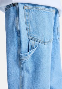 Jean en denim bleu clair avec un détail de poche visible et une couture contrastante jaune. Caractérisé par un tissu doux et texturé ainsi qu'une coupe décontractée.