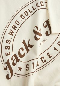 Cremefarvet bomulds t-shirt med et stort brunt rundt logo, der har teksten "Jack & J" og "SINCE 1980" i en prangende skrifttype.