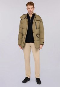 Giacca imbottita verde oliva con cappuccio, chiusura frontale con zip e tasche laterali. Dettaglio con zip nera sul petto, indossata sopra un top nero e pantaloni beige.