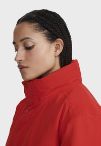Rote wasserdichte Jacke mit hohem Kragen und Druckknöpfen, mit glatter Textur und tailliertem Design, seitlich gezeigt.