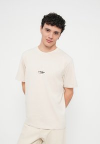 Light beige cotton t-shirt with short sleeves and a round neckline, featuring a small black "JJ Studios" logo on the chest.