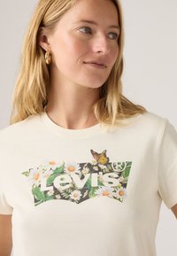 Κρεμ βαμβακερό μπλουζάκι με λουλουδένιο σχέδιο λογότυπου Levi's και μια πεταλούδα. Μανίκια κοντά και στρογγυλή λαιμόκοψη.