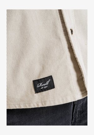 Beige corduroy shirt with vertical ribbing, featuring a black fabric label with white text reading “Reell est. 1997.” Bottom hem is stitched.