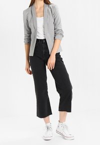Šedý pletený blazer s klopami, přes bílý top bez rukávů, doplněný o černé džíny s vysokým pasem a bílé tenisky. Slim fit s krátkým střihem.