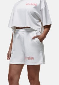 Maglietta bianca corta con logo rosa e pantaloni corti bianchi con stampa rosa; entrambi realizzati in morbido tessuto, con vita elastica e tasche.