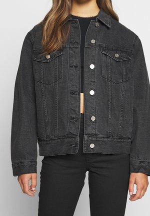 Vrouw draagt een zwarte kortgesneden spijkerjasje met zilveren knopen over een zwarte top en zwarte broek, staand tegen een effen achtergrond.