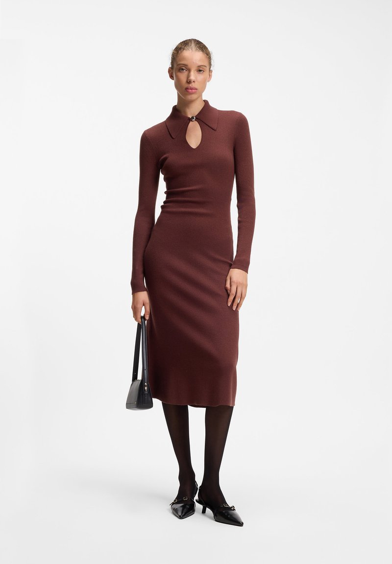 Femme vêtue d'une robe midi marron cintrée à manches longues avec col et détail œillet, des collants noirs, des talons noirs, et tenant un sac à main noir.